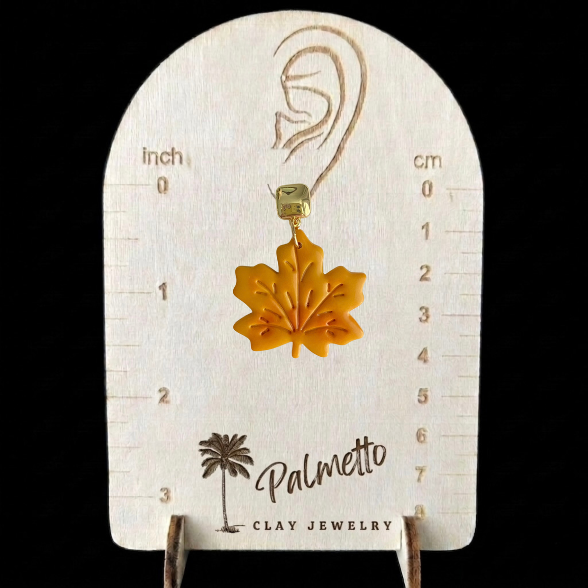Mustard polymer clay maple leaf earrings with embossed vein details and polished gold-tone posts on a Palmetto Clay Jewelry measurement tool.