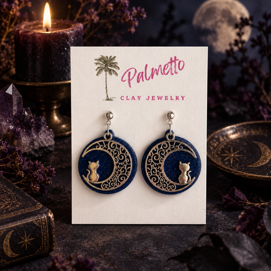 Midnight blue handmade earrings with ornate silver crescent moons and sitting cats on a Palmetto Clay Jewelry card.