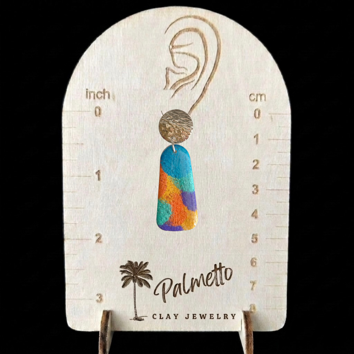 A pair of colorful hand-painted clay drop earrings displayed on a Palmetto Clay Jewelry measurement tool.