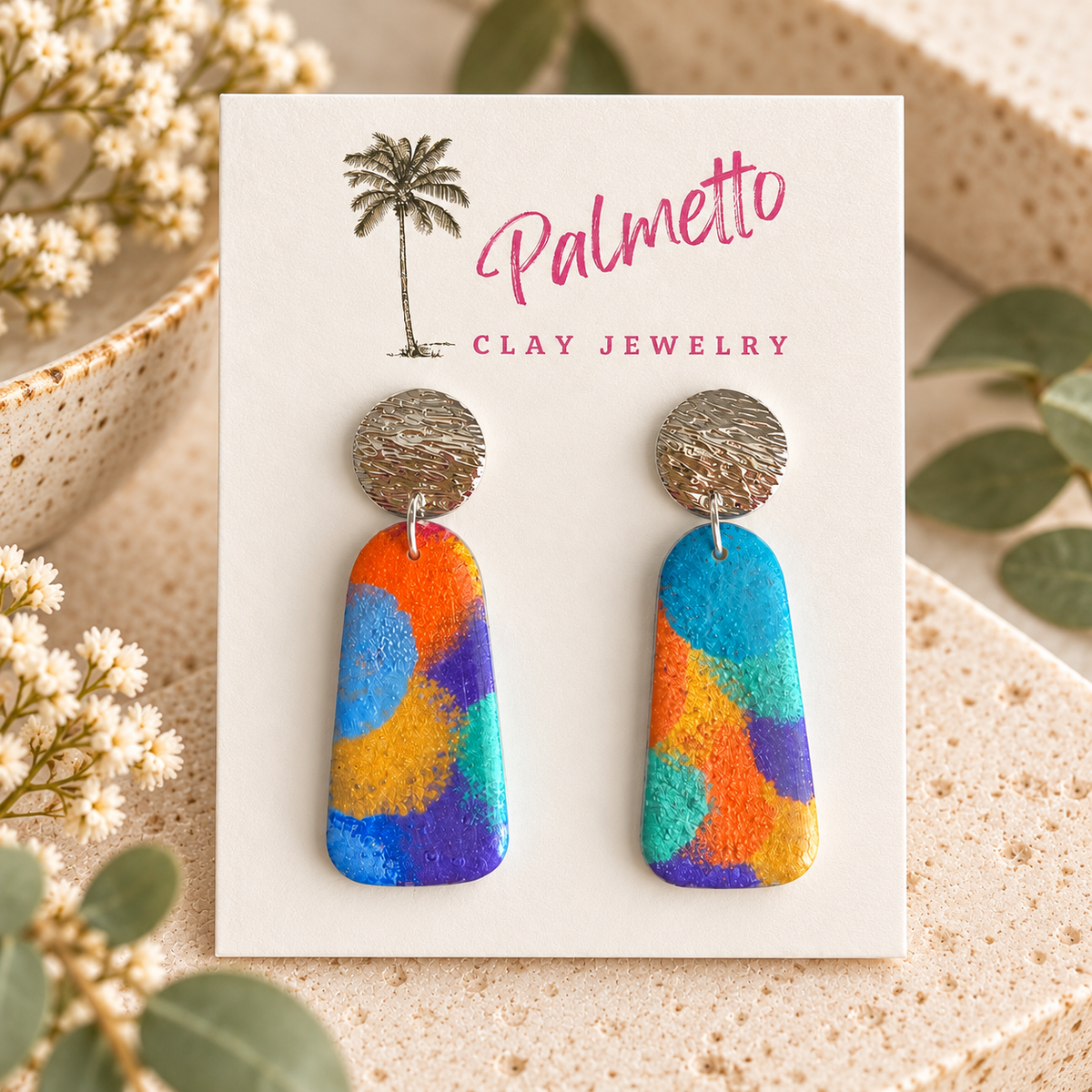 A pair of colorful hand-painted clay drop earrings displayed on a white Palmetto Clay Jewelry card.