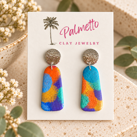 A pair of colorful hand-painted clay drop earrings displayed on a white Palmetto Clay Jewelry card.