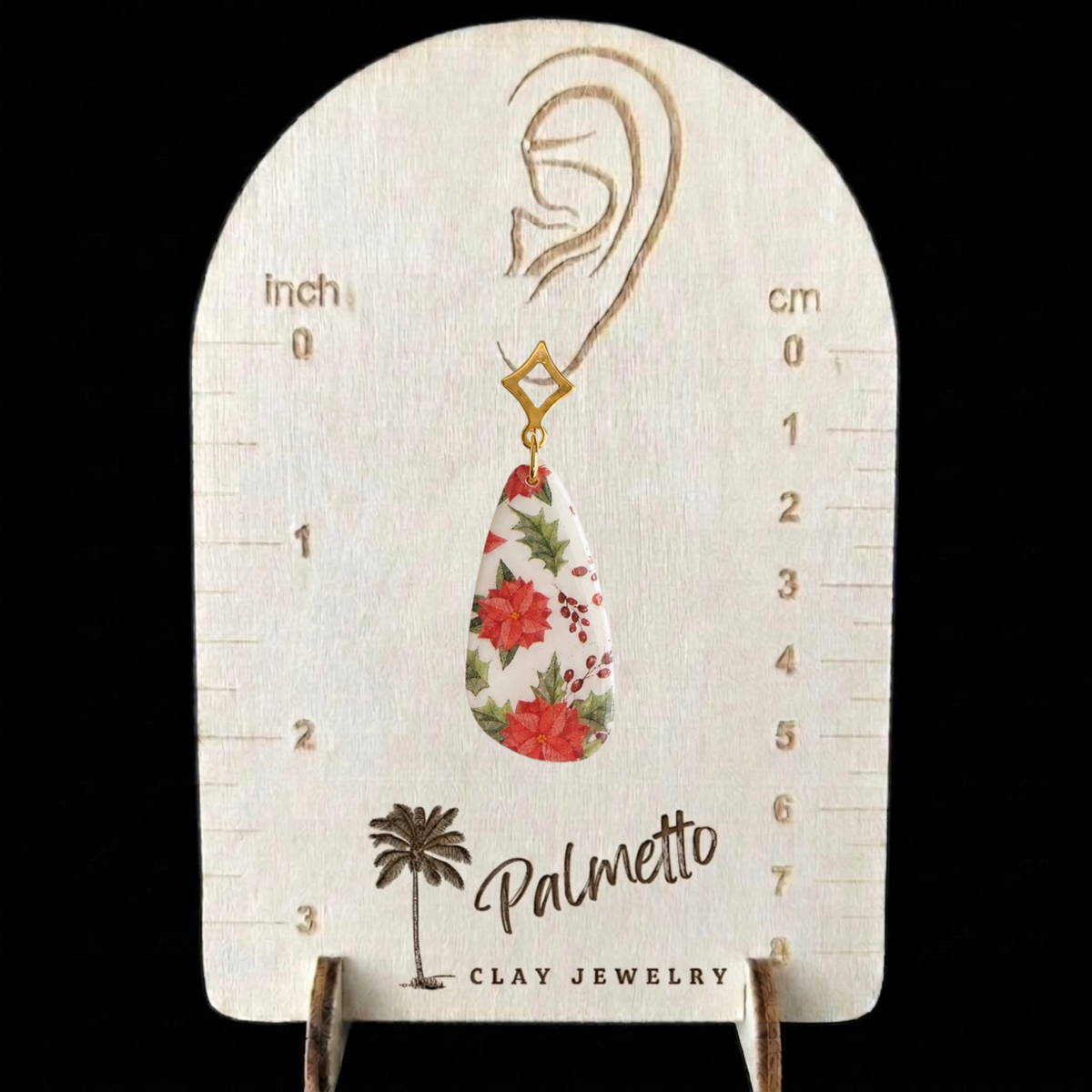 Handmade poinsettia Christmas earring with red flowers, green holly, berry details and gold geometric stud shown on an earring size measurement display.