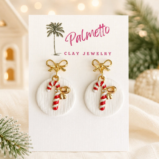 Handcrafted polymer clay Christmas earrings featuring red and white candy cane charms with gold bows on textured white circles and gold bow studs.