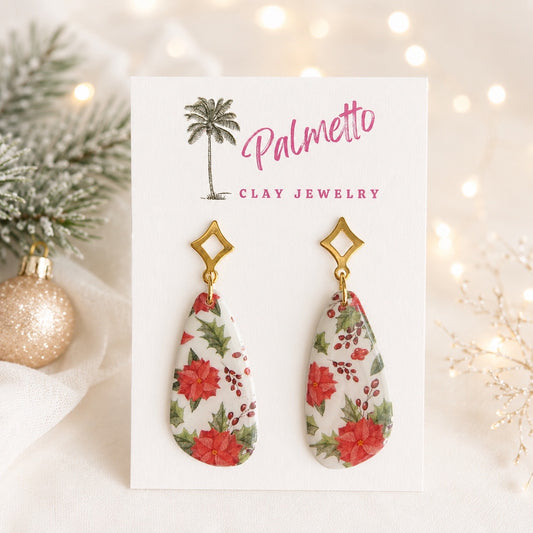 Handcrafted polymer clay Christmas earrings with red poinsettias, green holly leaves and berries on white teardrop shapes with gold-tone geometric studs.